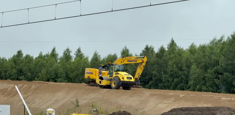 Żółta koparka gąsienicowa marki Komatsu oraz walec drogowy stojące na piaszczystym nasypie kolejowym na tle ściany lasu. U góry widoczne przewody sieci trakcyjnej. Maszyny budowlane na nasypie ziemnym w rejonie Głuszyna podczas prac przy budowie drugiego toru linii kolejowej nr 202. W tym rejonie doszło do tragicznego wypadku, w którym pod gąsienicami koparki zginęła 45-letnia pracownica