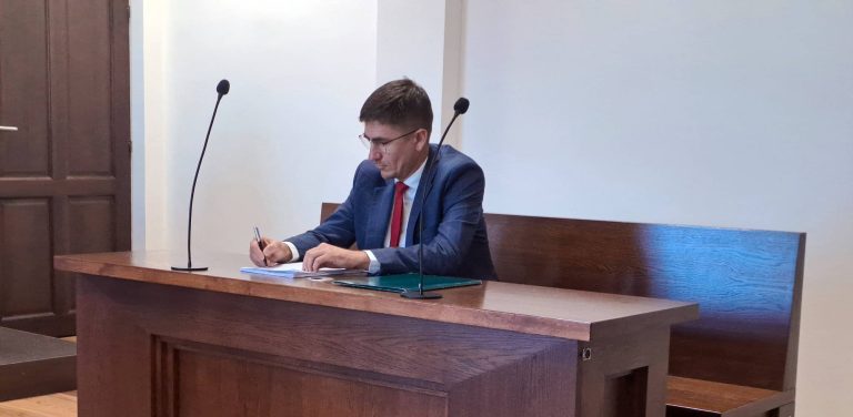 Mężczyzna w okularach, granatowym garniturze i czerwonym krawacie siedzi na sali sądowej przy drewnianym stole z mikrofonami i podpisuje dokumenty długopisem. To były burmistrz Debrzna Wojciech Kallas podczas procesu wytoczonego przez urząd miejski w sprawie zwrotu służbowego laptopa. Sąd rejonowy oddalił powództwo magistratu, uznając zniszczenie i utylizację sprzętu za skuteczne