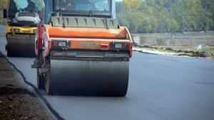 Pomarańczowy walec drogowy ubijający świeżą nawierzchnię asfaltową na budowanej trasie. W tle widoczna kolejna maszyna budowlana oraz pas zieleni. Gdański oddział GDDKiA uzyskał decyzję środowiskową dla ponad 22-kilometrowej obwodnicy Człuchowa