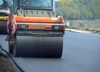 Pomarańczowy walec drogowy ubijający świeżą nawierzchnię asfaltową na budowanej trasie. W tle widoczna kolejna maszyna budowlana oraz pas zieleni. Gdański oddział GDDKiA uzyskał decyzję środowiskową dla ponad 22-kilometrowej obwodnicy Człuchowa
