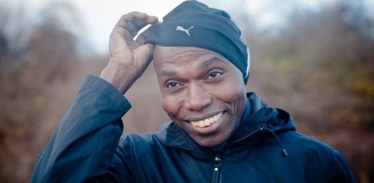 Uśmiechnięty Wilson Kipketer w czarnej sportowej kurtce i czarnej czapce marki Puma, którą lekko unosi dłonią