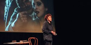 Aktorka Beata Fudalej w ciemnym stroju i okularach przemawia na scenie. W tle widać duży ekran z kadrem filmowym przedstawiającym mężczyznę celującego z pistoletu oraz kobietę, a z boku stolik ze stosem książek Stanisława Ignacego Witkiewicza