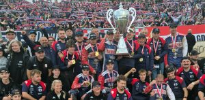 Żużlowcy i sztab Wybrzeża Gdańsk ze złotymi medalami na szyjach unoszą okazały srebrny puchar mistrzowski. W tle widać wypełnione trybuny z kibicami unoszącymi w górę klubowe szaliki