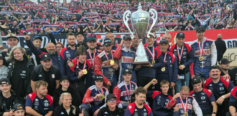 Żużlowcy i sztab Wybrzeża Gdańsk ze złotymi medalami na szyjach unoszą okazały srebrny puchar mistrzowski. W tle widać wypełnione trybuny z kibicami unoszącymi w górę klubowe szaliki