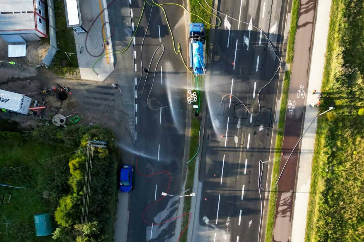 Widok z drona prostopadle z góry na zablokowaną, wielopasmową jezdnię miejską. Na drodze stoją czerwone wozy strażackie i pojazdy techniczne, po asfalcie poprowadzone są węże strażackie, a nad ulicą unoszą się strumienie mgły z kurtyn wodnych