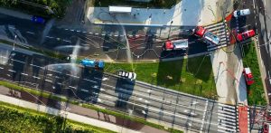 Widok z drona prostopadle z góry na zablokowaną, wielopasmową jezdnię miejską. Na drodze stoją czerwone wozy strażackie i pojazdy techniczne, po asfalcie poprowadzone są węże strażackie, a nad ulicą unoszą się strumienie mgły z kurtyn wodnych