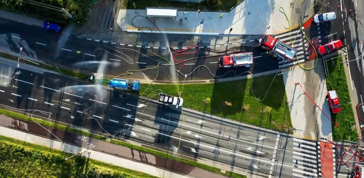 Widok z drona prostopadle z góry na zablokowaną, wielopasmową jezdnię miejską. Na drodze stoją czerwone wozy strażackie i pojazdy techniczne, po asfalcie poprowadzone są węże strażackie, a nad ulicą unoszą się strumienie mgły z kurtyn wodnych