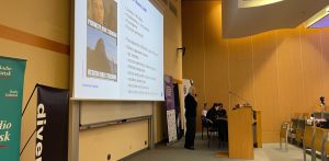 Prelekcja w auli Uniwersytetu Gdańskiego podczas konferencji naukowej z okazji Światowego Dnia Zapobiegania Samobójstwom. Kobieta z mikrofonem przemawia w auli uniwersyteckiej obok mównicy i dużego ekranu z prezentacją na temat trudności psychicznych studentów. Po lewej stronie widoczne są banery Radia Gdańsk i DiversityPL