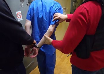 Dwóch policjantów prowadzi korytarzem skutego kajdankami mężczyznę w niebieskim kombinezonie. Jeden z funkcjonariuszy ma na sobie kamizelkę taktyczną z białym napisem "policja". 46-latek miał podłożyć substancję łatwopalną przy jednych film w Elblągu