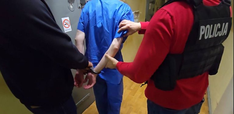 Dwóch policjantów prowadzi korytarzem skutego kajdankami mężczyznę w niebieskim kombinezonie. Jeden z funkcjonariuszy ma na sobie kamizelkę taktyczną z białym napisem "policja". 46-latek miał podłożyć substancję łatwopalną przy jednych film w Elblągu