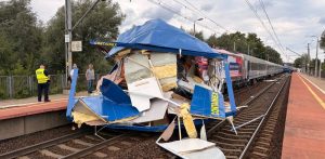 Zmiażdżona część naczepy TIR-a wbita w przód lokomotywy pociągu pasażerskiego PKP Intercity stojącego na torach przy peronie. Wokół leżą połamane elementy konstrukcji i wełna izolacyjna, a na peronie stoją strażak oraz świadkowie zdarzenia. Do zderzenia TIR-a z pociągiem doszło na przejeździe nieopodal przystanku Gdańsk Lipce