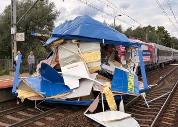 Zmiażdżona część naczepy TIR-a wbita w przód lokomotywy pociągu pasażerskiego PKP Intercity stojącego na torach przy peronie. Wokół leżą połamane elementy konstrukcji i wełna izolacyjna, a na peronie stoją strażak oraz świadkowie zdarzenia. Do zderzenia TIR-a z pociągiem doszło na przejeździe nieopodal przystanku Gdańsk Lipce