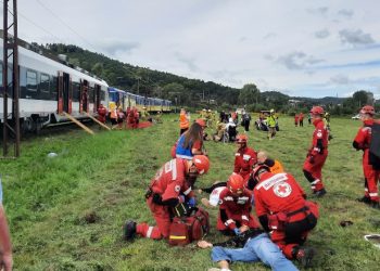 Ratownicy w czerwonych kombinezonach i kaskach udzielają pierwszej pomocy osobie leżącej na trawie, na tle stojącego na torach pociągu pasażerskiego. Wokół na łące widać liczne służby ratunkowe, ewakuację ze składu oraz sprzęt medyczny. Czołowe zderzenie dwóch pociągów w Gdyni było elementem ćwiczeń w ramach manewrów ratowniczych Pomerania - Reda 2026