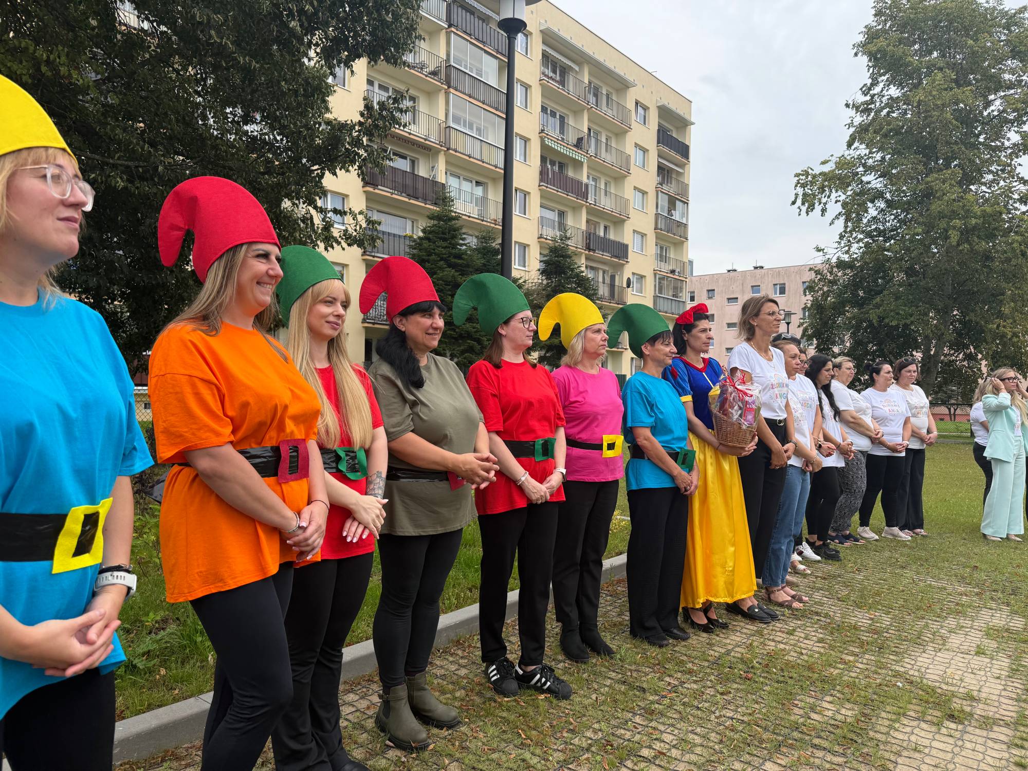 Pracownice Żłobka Miejskiego nr 1 w Słupsku stojące w rzędzie na trawniku na tle bloku mieszkalnego. Opiekunki są przebrane w kolorowe stroje i wysokie czapki krasnoludków oraz suknię Królewny Śnieżki z koszykiem w ręku