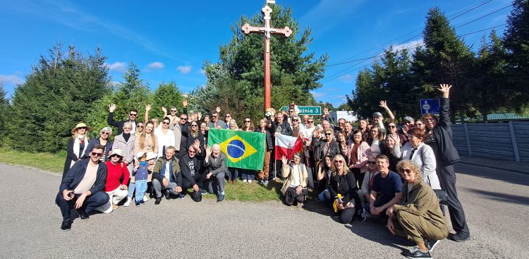 Duża grupa uśmiechniętych ludzi w różnym wieku pozuje na poboczu drogi pod wysokim, drewnianym krzyżem przydrożnym. W centrum uczestnicy trzymają rozłożoną flagę Brazylii oraz flagę Polski, a część osób macha w stronę aparatu. W tle widać zielone drzewa i bezchmurne niebo. Potomkowie polskich emigrantów z Brazylii z flagami obu ojczyzn przy przydrożnym krzyżu w Łęgu pod Czerskiem, który przed laty postawili ich przodkowie.
