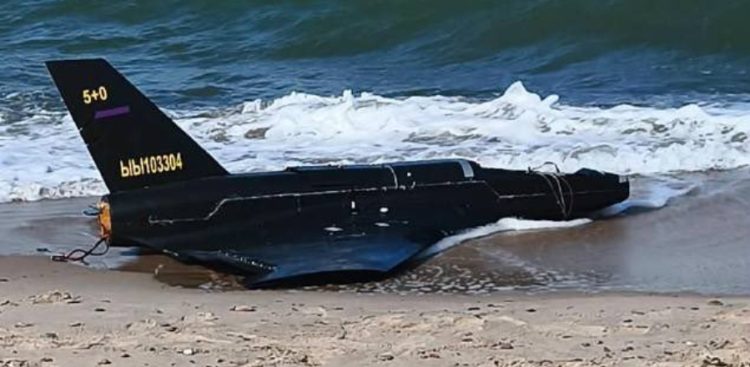 Dron na plaży w Rusinowie koło Jarosławca. Czarny wojskowy dron bezzałogowy leżący na piaszczystej plaży u brzegu morza, częściowo omywany przez spienione fale. Na stateczniku pionowym widoczne są żółte oznaczenia alfanumeryczne