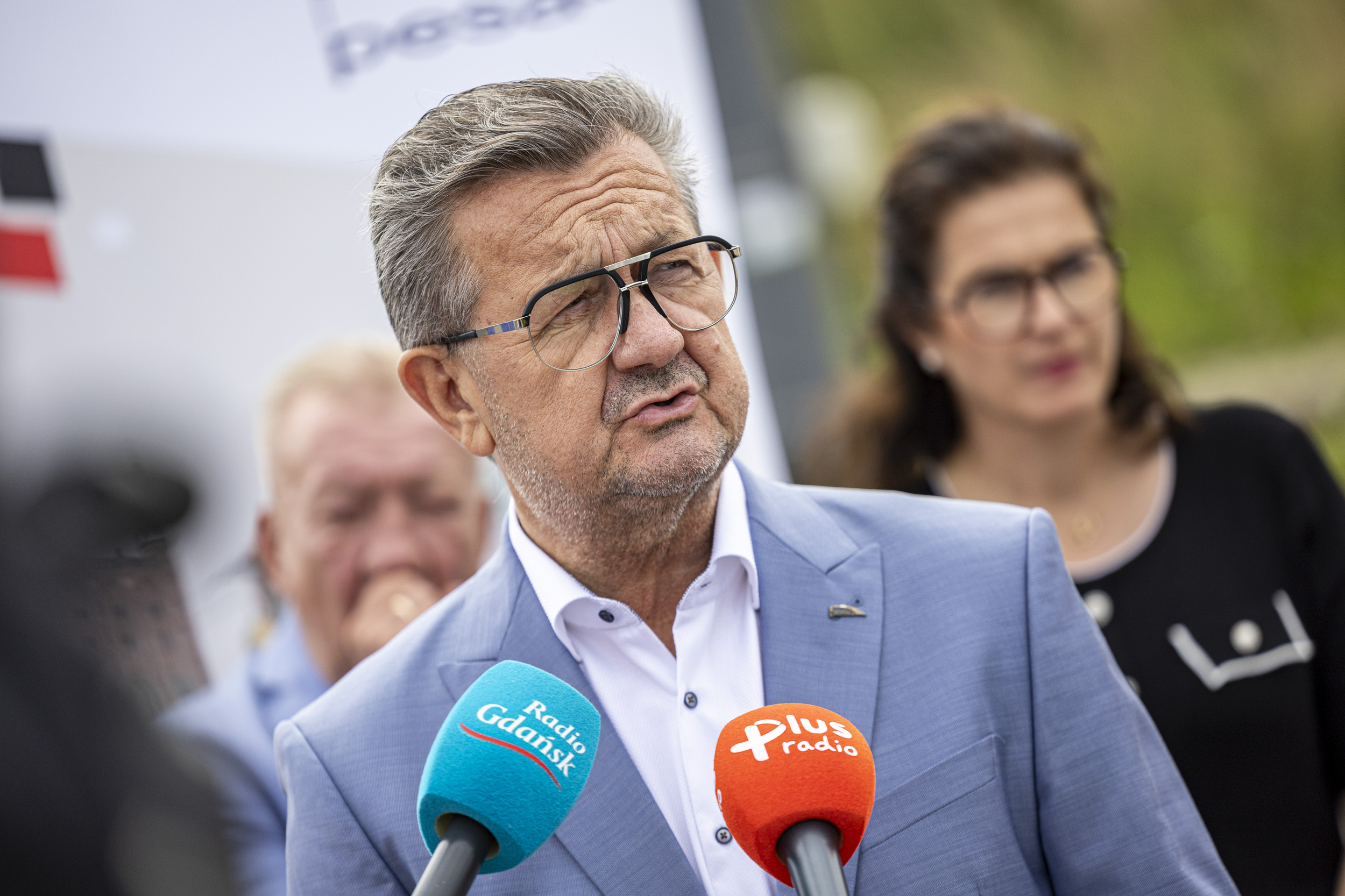 Zbliżenie na Krzysztofa Zdziarskiego, prezesa firmy Pesa Bydgoszcz, ubranego w jasnoniebieską marynarkę i okulary, przemawiającego do mikrofonów z logo Radia Gdańsk. W nieostrym tle widoczna jest prezydent Aleksandra Dulkiewicz