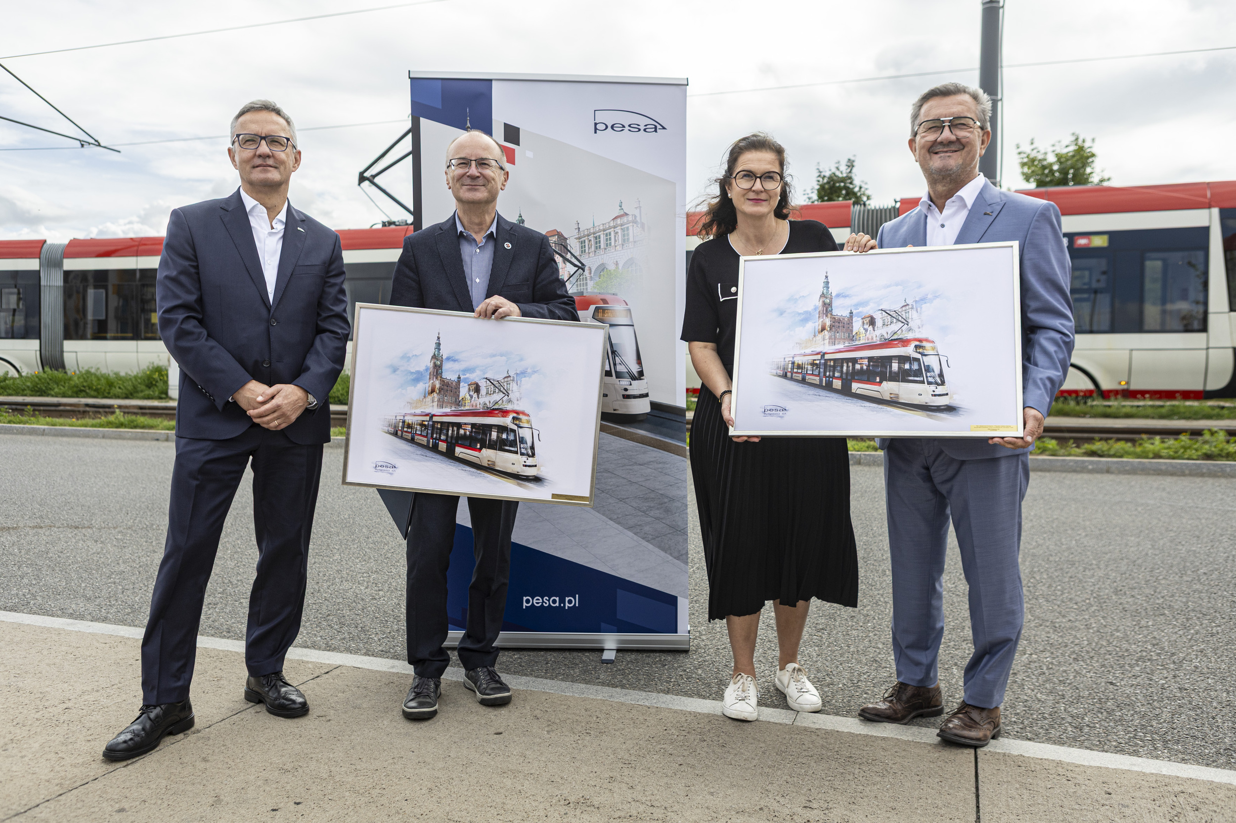 Cztery osoby - w tym prezydent Gdańska Aleksandra Dulkiewicz oraz Krzysztof Zdziarski, prezes zarządu i dyrektor zarządzający spółki Pesa Bydgoszcz - w eleganckich strojach stoją na tle tramwaju i baneru PESA, trzymając w rękach dwie oprawione w ramy grafiki przedstawiające nowy tramwaj na tle gdańskiego Ratusza Głównego Miasta
