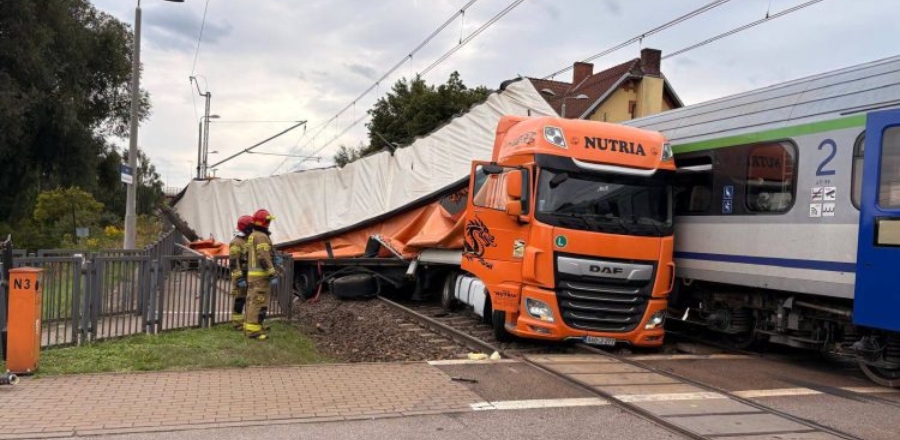Pomarańczowy ciągnik siodłowy marki DAF ze zniszczoną, zmiażdżoną naczepą stojący na przejeździe kolejowym tuż obok wagonu pociągu pasażerskiego. W tle widać przewróconą konstrukcję naczepy, a z lewej strony dwóch strażaków prowadzących działania na torowisku. TIR zderzył się z pociągiem nieopodal stacji Gdańsk Lipce. 42-letni kierowca ciężarówki usłyszał zarzut sprowadzenia bezpośredniego niebezpieczeństwa katastrofy w ruchu lądowym