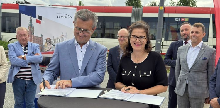 Prezydent Gdańska Aleksandra Dulkiewicz oraz prezes zarządu PESA Bydgoszcz Krzysztof Zdziarski podpisują dokumenty przy stoliku na świeżym powietrzu. W tle stoją obserwatorzy, baner z logo Pesa oraz nowoczesny tramwaj. Gdańsk będzie mieć najdłuższe tramwaje w Polsce
