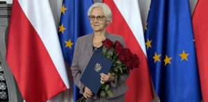 Uśmiechnięta Danuta Wałęsa w okularach i szarym kostiumie trzymająca bukiet czerwonych róż oraz granatową teczkę z godłem Polski na tle flag Polski i Unii Europejskiej. Danuta Wałęsa rozpoczynająca działanie dla Pomorskiej Wojewódzkiej Rady Konsultacyjnej do spraw Działaczy Opozycji Antykomunistycznej i Osób Represjonowanych z Powodów Politycznych
