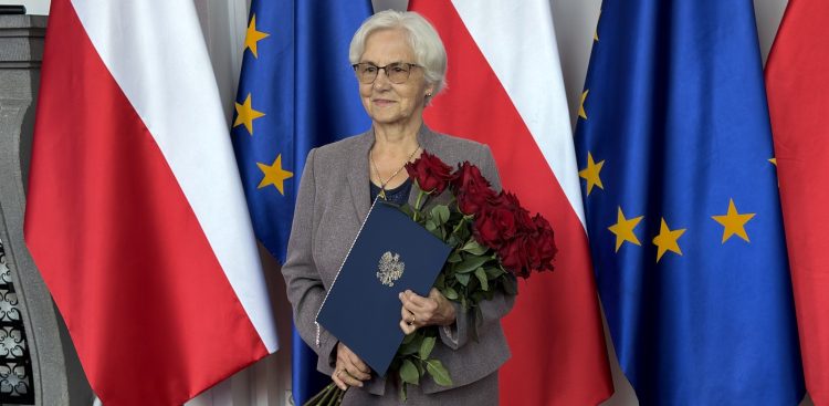 Uśmiechnięta Danuta Wałęsa w okularach i szarym kostiumie trzymająca bukiet czerwonych róż oraz granatową teczkę z godłem Polski na tle flag Polski i Unii Europejskiej. Danuta Wałęsa rozpoczynająca działanie dla Pomorskiej Wojewódzkiej Rady Konsultacyjnej do spraw Działaczy Opozycji Antykomunistycznej i Osób Represjonowanych z Powodów Politycznych