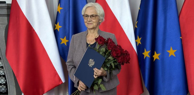 Uśmiechnięta Danuta Wałęsa w okularach i szarym kostiumie trzymająca bukiet czerwonych róż oraz granatową teczkę z godłem Polski na tle flag Polski i Unii Europejskiej. Danuta Wałęsa rozpoczynająca działanie dla Pomorskiej Wojewódzkiej Rady Konsultacyjnej do spraw Działaczy Opozycji Antykomunistycznej i Osób Represjonowanych z Powodów Politycznych