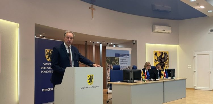 Marszałek województwa pomorskiego Mieczysław Struk przemawia przy mównicy z herbem województwa podczas konferencji prasowej w Urzędzie Marszałkowskim w Gdańsku. W tle widoczny jest stół prezydialny oraz banery promocyjne Funduszy Europejskich dla Pomorza