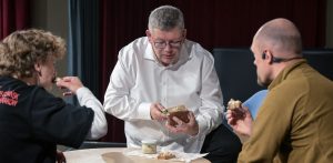 Trzy osoby siedzą przy stole i jedzą chleb. Mężczyzna w białej koszuli i okularach smaruje nożem kromkę pieczywa pastą ze słoika, a pozostali dwaj mężczyźni trzymają w dłoniach przygotowane kromki. Degustacja tradycyjnych pomorskich przysmaków podczas nagrywania materiału o dawnej kuchni Gdańska. Uczestnicy spotkania próbują chleba ze smalcem ze słoika, przybliżając zapomniane lokalne receptury