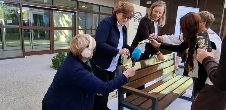Grupa osób wspólnie maluje pędzlami drewnianą ławkę na żółty kolor przed budynkiem Centrum Opieki Wytchnieniowej; jedna z kobiet (Anna Rębas z Radia Gdańsk) trzyma mikrofon radiowy, a inna osoba nagrywa akcję smartfonem