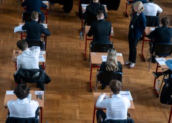 Na zdjęciu znajdują się maturzyści piszący egzamin. Wszyscy są w białych koszulach i siedzą na drewnianych ławkach, na drewnianej auli.