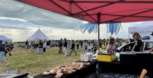 Plenerowy piknik na lotnisku. Na pierwszym planie stoisko pod różowym zadaszeniem z przekąskami, w tle duży biały namiot i spacerujący ludzie.