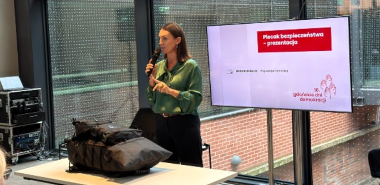 Debata w czasie Gdańskich Dni Demokracji. Beata Rutkiewicz, wojewoda pomorska, prezentuje plecak ewakuacyjny. Przed nią stoi biurko, za nią ekran z prezentacją