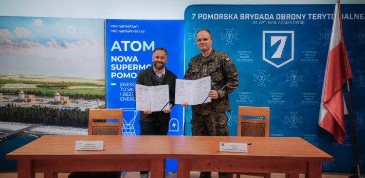 Przedstawiciel spółki Polskie Elektrownie Jądrowe i dowódca 7. Pomorskiej Brygady Obrony Terytorialnej stoją za stołem i prezentują podpisany dokument. W tle widać baner informacyjny o energii jądrowej na Pomorzu oraz ściankę 7PBOT z flagą Polski