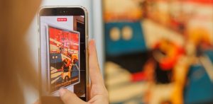 Osoba trzyma telefon komórkowy i nagrywa film przedstawiający plakat 51. Festiwalu Polskich Filmów Fabularnych z motywem statku. W tle widać rozmyty fragment tego samego, kolorowego plakatu w większym formacie
