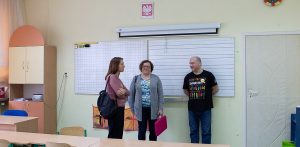 Nauczyciele w szkolnej klasie. Dwie kobiety i mężczyzna rozmawiają w sali lekcyjnej na tle białych tablic i wiszącego nad nimi godła Polski