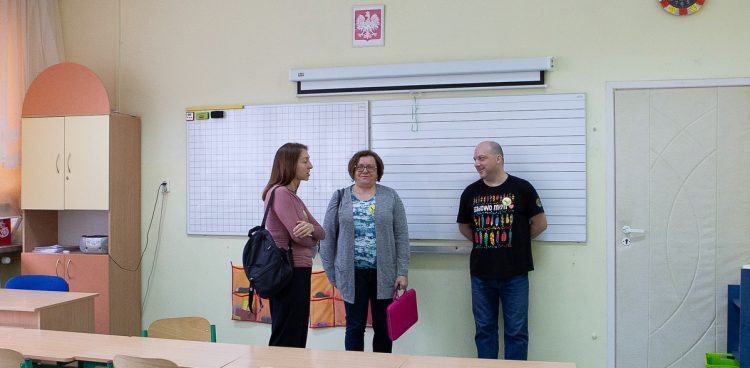Nauczyciele w szkolnej klasie. Dwie kobiety i mężczyzna rozmawiają w sali lekcyjnej na tle białych tablic i wiszącego nad nimi godła Polski