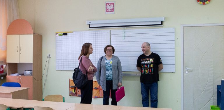 Nauczyciele w szkolnej klasie. Dwie kobiety i mężczyzna rozmawiają w sali lekcyjnej na tle białych tablic i wiszącego nad nimi godła Polski