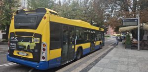 Żółto-niebieski autobus miejski Scania Citywide (MZK Słupsk, numer boczny 3066) stojący na przystanku w centrum miasta. W tle widać elektroniczną tablicę odjazdów, zieleń miejską oraz ogródek kawiarniany. Zarząd Infrastruktury Miejskiej w Słupsku wprowadził nowe bilety, a także zmiany w istniejących. Mają one zachęcać mieszkańców do częstszego korzystania z komunikacji zbiorowej