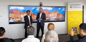 Na zdjęciu znajduje się dwóch mężczyzn na tle zdjęcia panoramy Gdańska. Przed nimi siedzą tyłem ludzie, robią im zdjęcia telefonami.