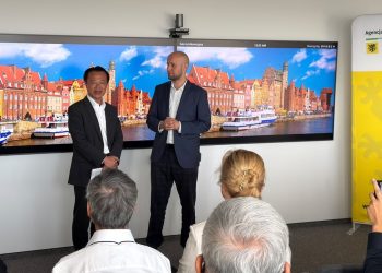 Na zdjęciu znajduje się dwóch mężczyzn na tle zdjęcia panoramy Gdańska. Przed nimi siedzą tyłem ludzie, robią im zdjęcia telefonami.