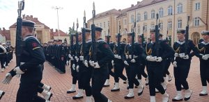 Uroczysta przysięga wojskowa w Gdyni. Grupa podchorążych w galowych mundurach marynarskich, białych czapkach i getrach maszeruje w szyku z bronią na placu apelowym przed budynkiem uczelni