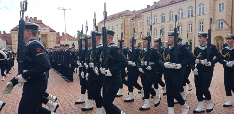 Uroczysta przysięga wojskowa w Gdyni. Grupa podchorążych w galowych mundurach marynarskich, białych czapkach i getrach maszeruje w szyku z bronią na placu apelowym przed budynkiem uczelni