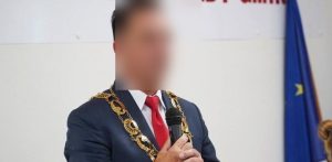 Na zdjęciu znajduje się mężczyzna w marynarce i oficjalnym łańcuchu prezydenta miasta. Jego twarz jest zamazana. W tle flaga Unii Europejskiej