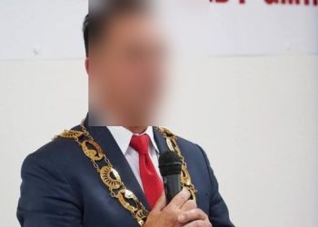 Na zdjęciu znajduje się mężczyzna w marynarce i oficjalnym łańcuchu prezydenta miasta. Jego twarz jest zamazana. W tle flaga Unii Europejskiej