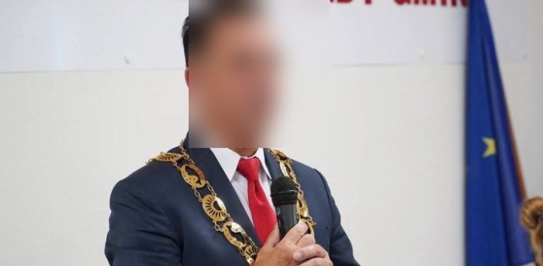 Na zdjęciu znajduje się mężczyzna w marynarce i oficjalnym łańcuchu prezydenta miasta. Jego twarz jest zamazana. W tle flaga Unii Europejskiej