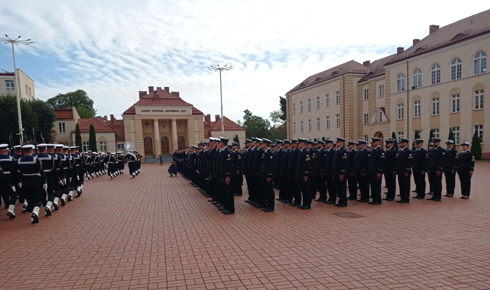 Szeroki kadr placu apelowego: po prawej stronie podchorążowie stoją na baczność w równym szyku, a po lewej maszeruje kolumna marynarzy z orkiestrą na tle gmachu z napisem "Amor Patriae Suprema Lex"