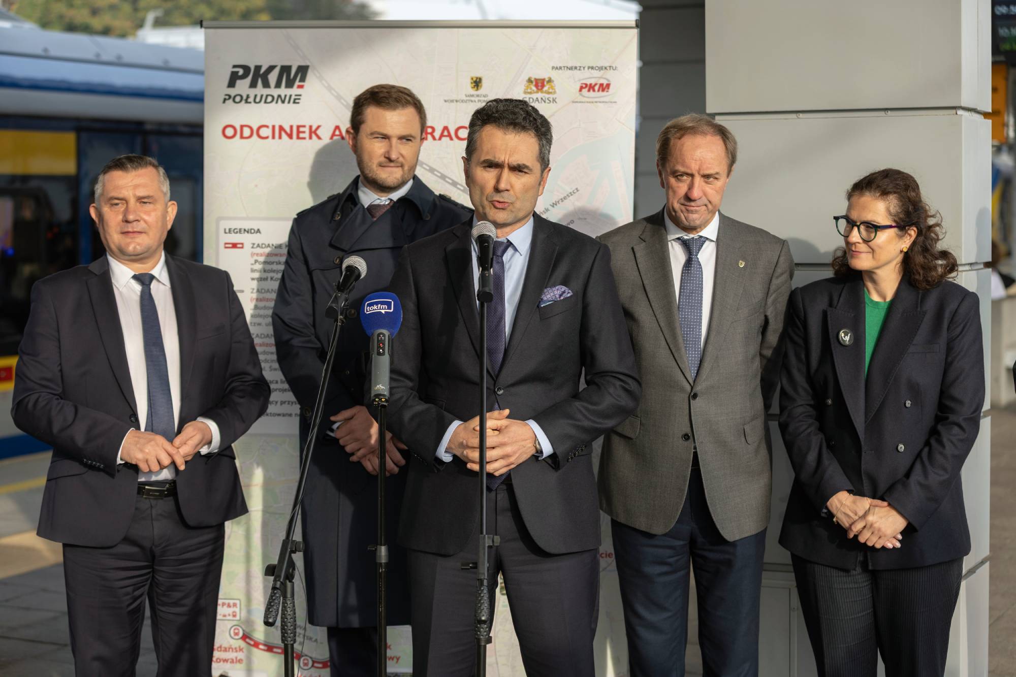 Wiceminister infrastruktury Piotr Malepszak przemawia do mikrofonów na tle planszy informacyjnej PKM Południe, w otoczeniu Grzegorza Mocarskiego, Piotra Borawskiego, Mieczysława Struka i Aleksandry Dulkiewicz