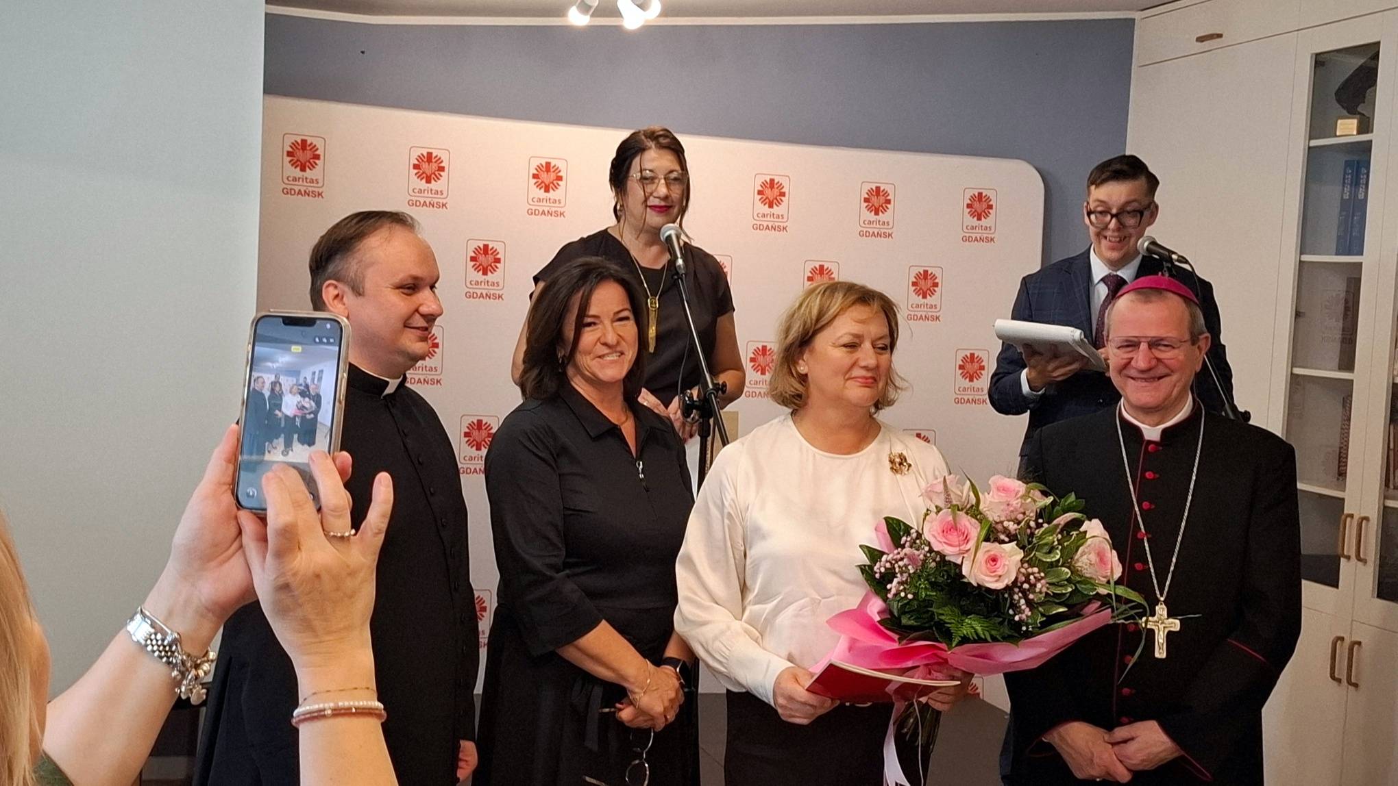 Uczestnicy uroczystości na tle ścianki z logo Caritas. w centrum dyrektor placówki trzyma bukiet róż, obok stoją abp Tadeusz Wojda. Na pierwszym planie widać dłoń robiącą im zdjęcie smartfonem