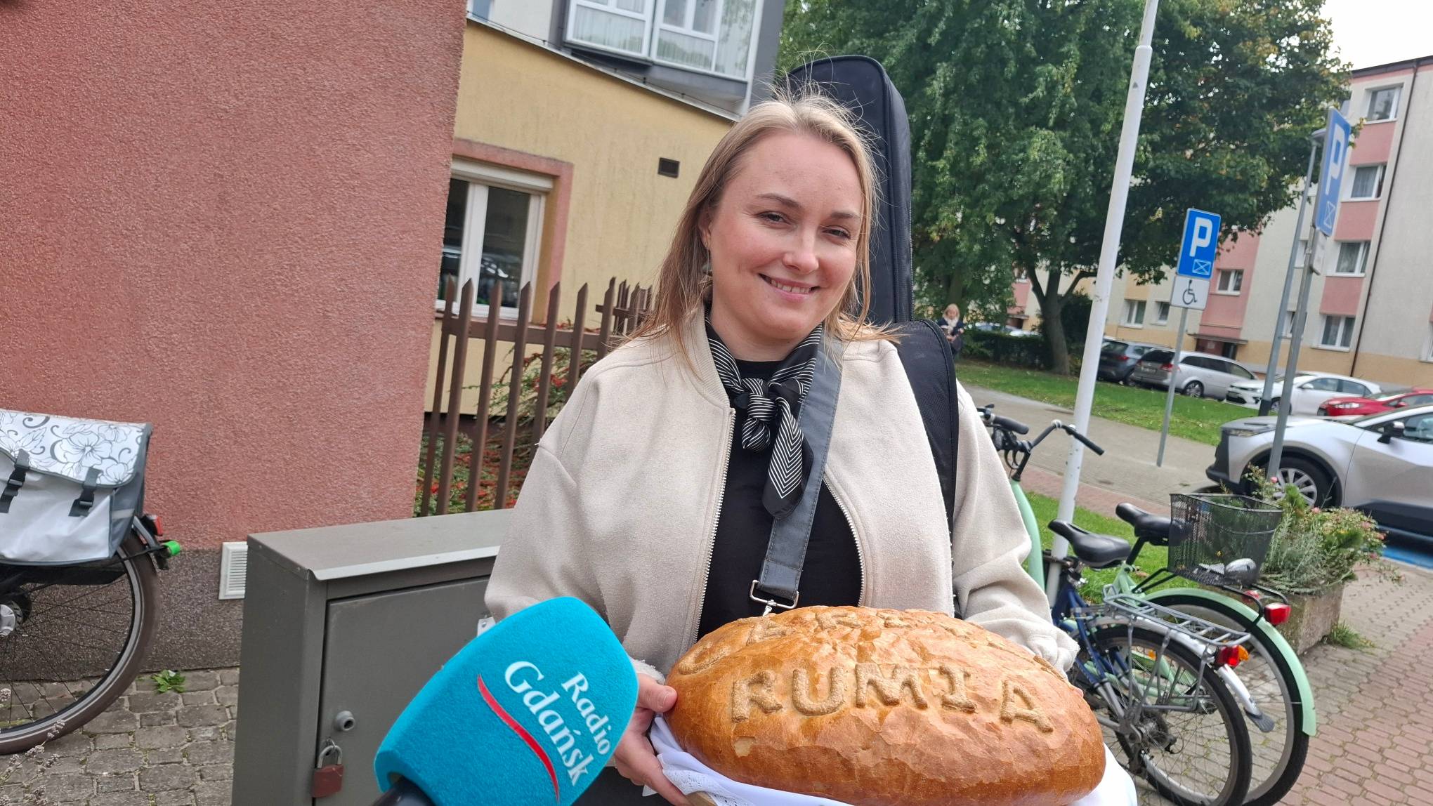Uśmiechnięta kobieta z pokrowcem na instrument na plecach trzyma duży bochenek chleba z wypieczonym napisem "RUMIA", stojąc przed mikrofonem Radia Gdańsk na tle budynku